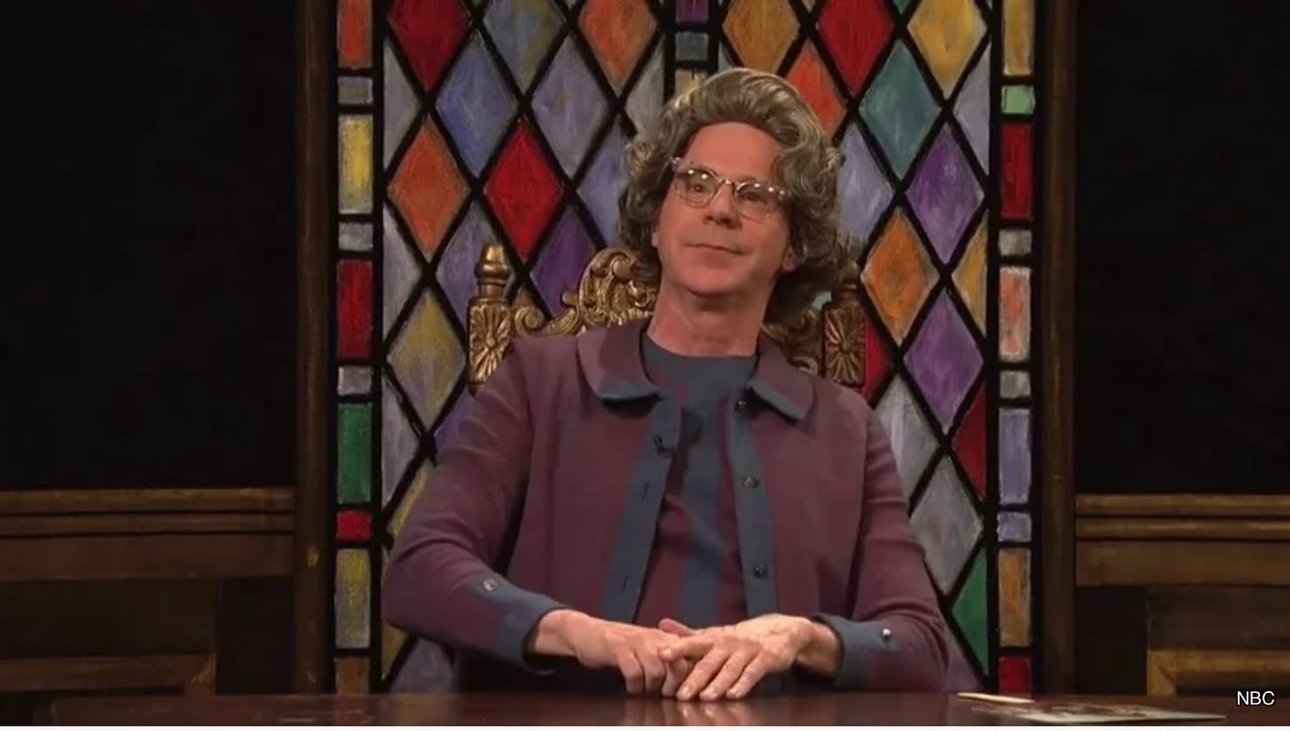 Actor and comedian Dana Carvey pictured here as his Saturday Night Live character, Enid Strict, with a gray-and-white-haired wig on, cat-eye glasses, dressed in a purple suit with navy blue accents, and seated at a desk in front of a multi-colored stained glass window