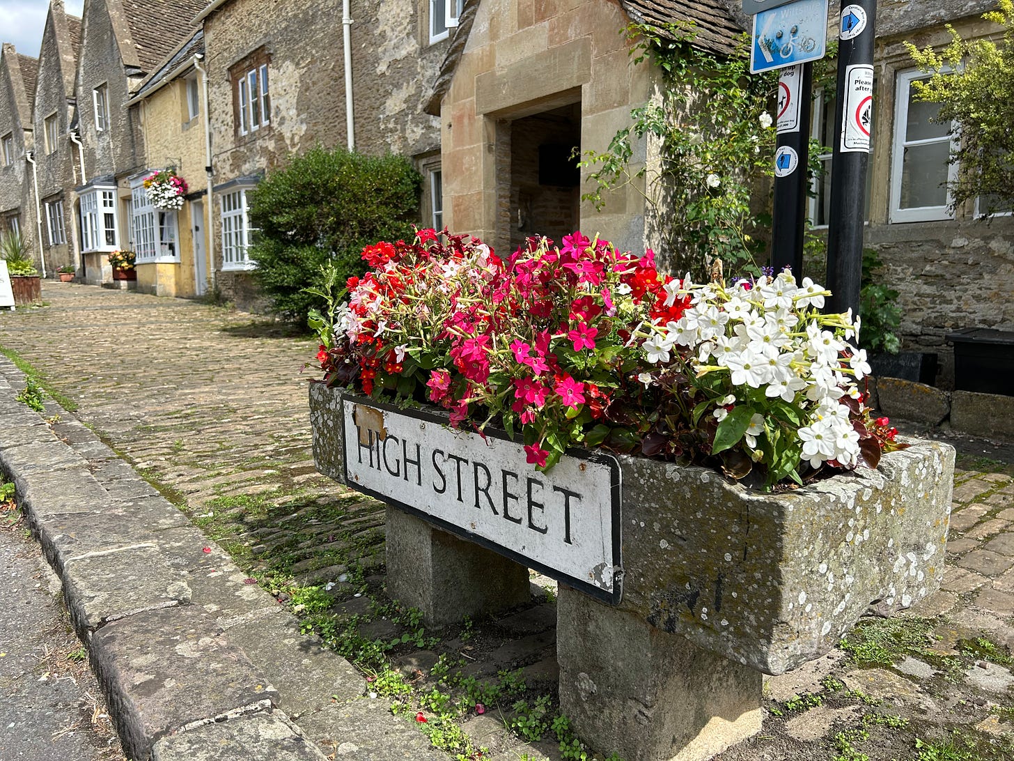 A street sign for the High Street on the side of a stone flower trough filled with summer flowers. Behind is a row of stone cottages and a cobbled pavement. Image: Roland's Travels The High Street, Corsham, Wiltshire