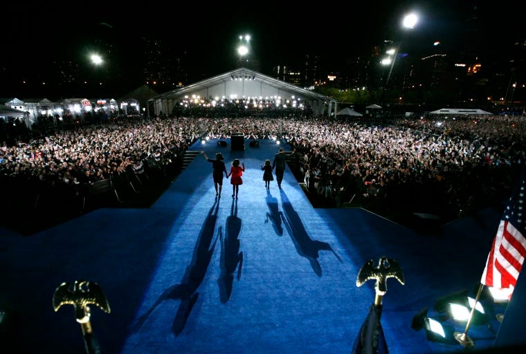 This image shows a 4 November 2008 photo of the Obama family--President-Elect Barak Obama (far right), First Lady Michelle Obama (far left), Malia Obama (second left), and Sasha Obama (second right)--as they walk onto the stage in Chicago, Illinois's Grant Park to celebrate Obama's presidential-election victory. This photo is taken from behind the Obamas as they walk toward the stage's front area. This image shows a 4 November 2008 photo of the Obama family--President-Elect Barak Obama (far right), First Lady Michelle Obama (far left), Malia Obama (second left), and Sasha Obama (second right)--as they walk onto the stage in Chicago, Illinois's Grant Park to celebrate Obama's presidential-election victory. This photo is taken from behind the Obamas as they walk toward the stage's front area.