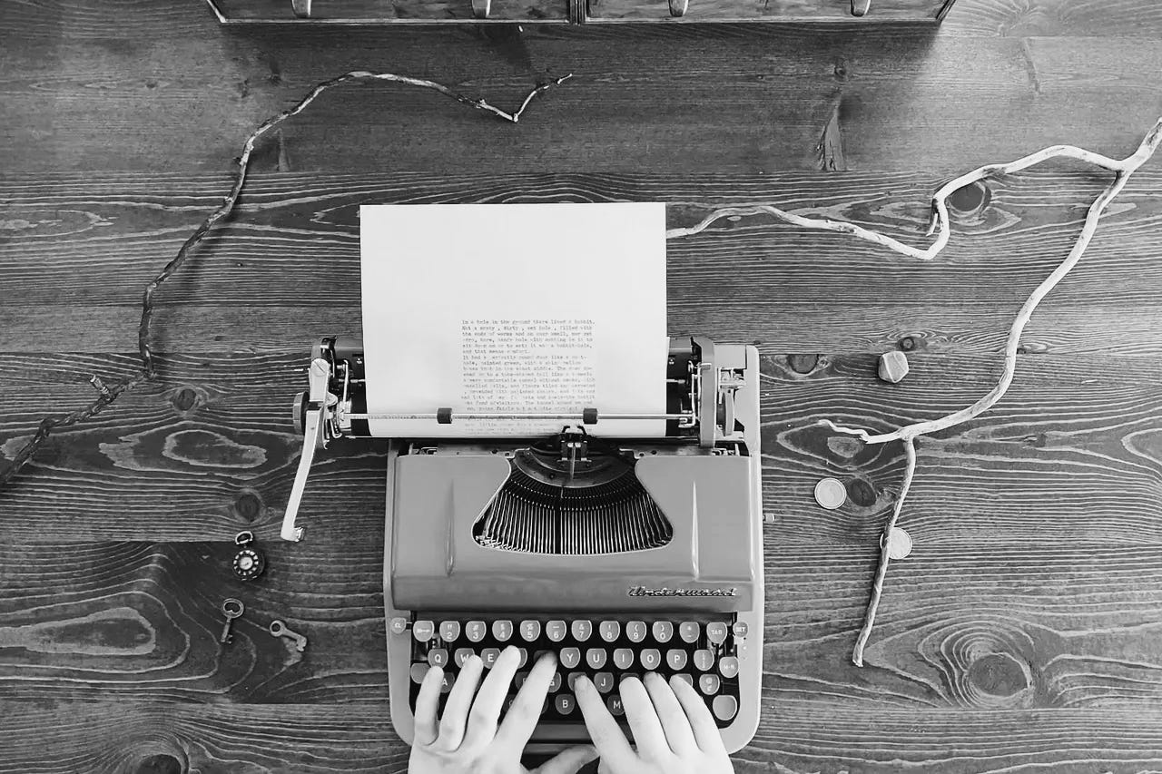 Black and white photograph of a typewriter on a wooden desk. My pale hands are typing.