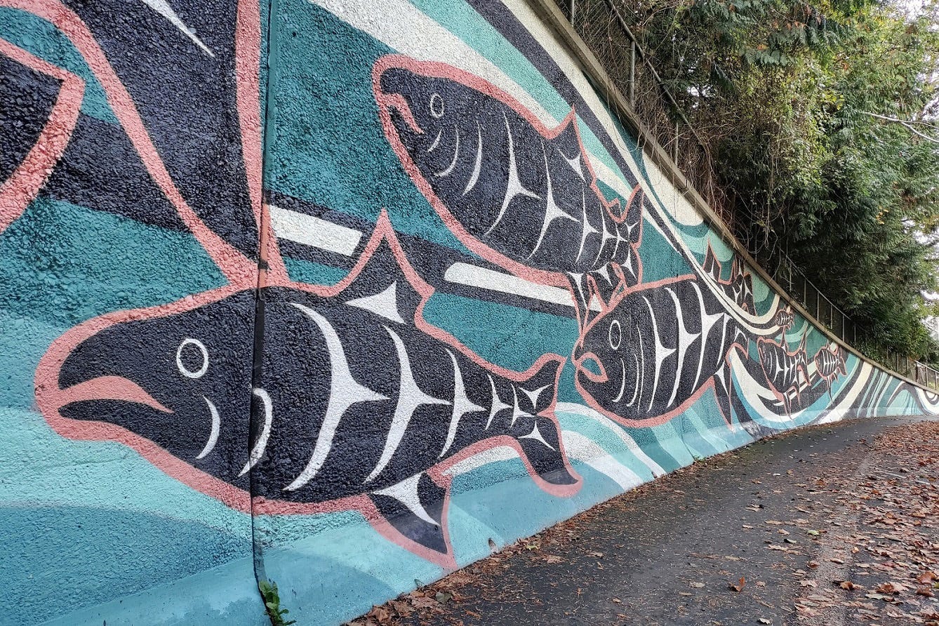 An outdoor infrastructure wall on a street is covered in a mural by Jason LaClair, an indigenous artist. It depicts a stylized blue river background with black and white salmon swimming in it, outlined in red.
