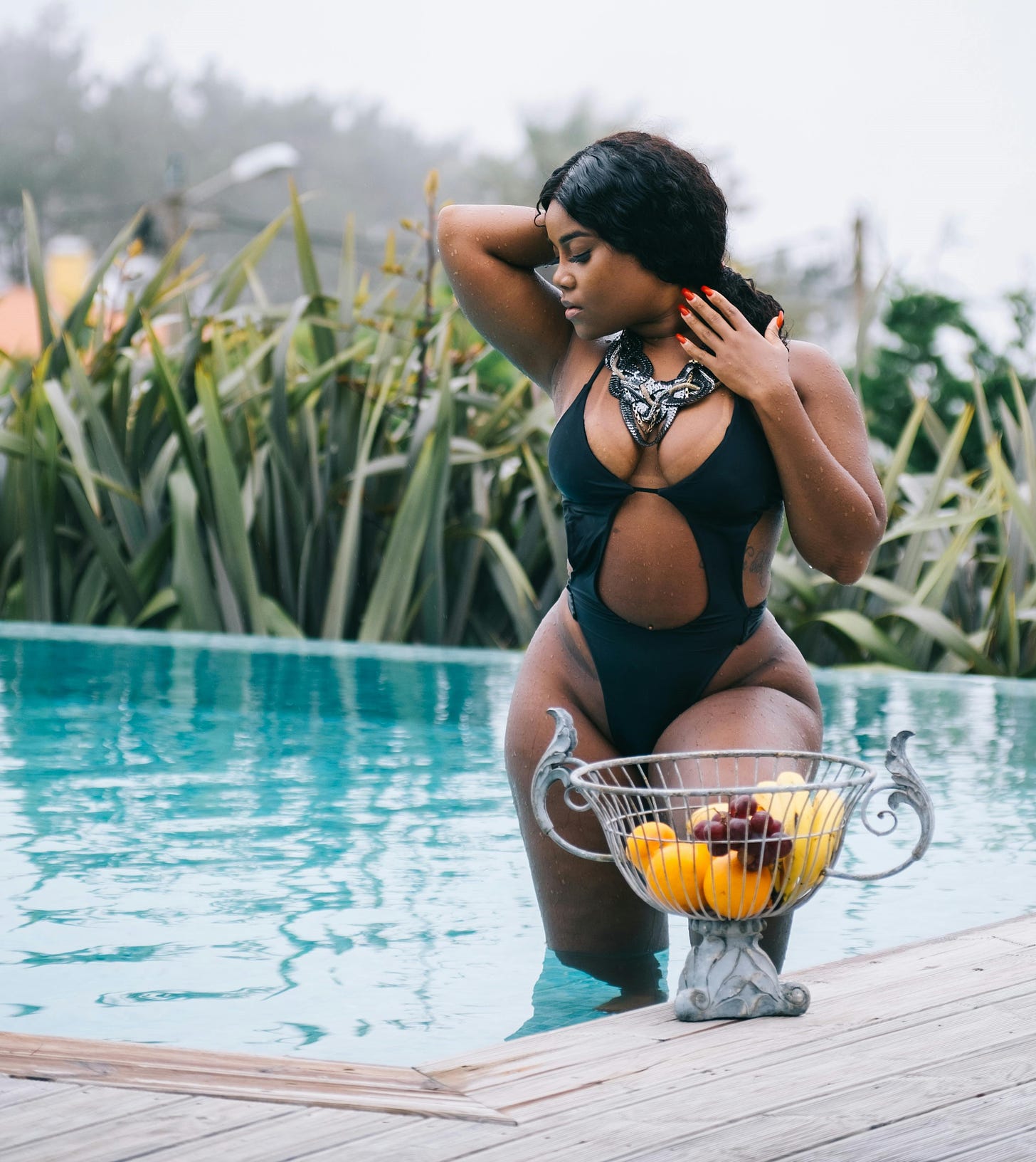 Woman, dark, black hair, in black one-piece bathing suit in pool with bowl of fruit in foreground.