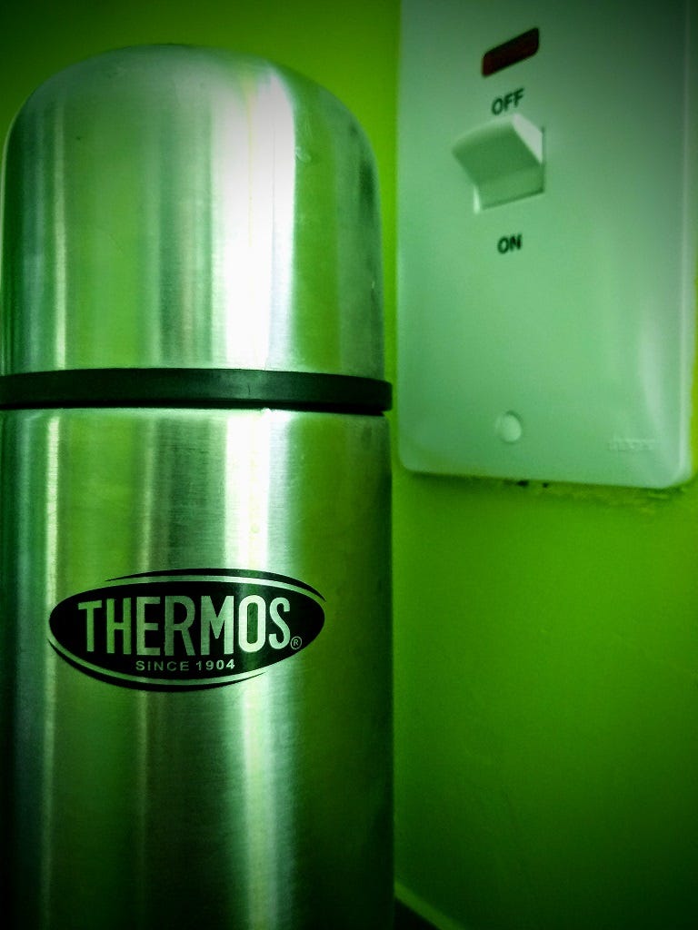 An unopened large silver vacuum flask stands bathed in sickly green light next to a large wall-mounted electrical switch set to the 'OFF' position.