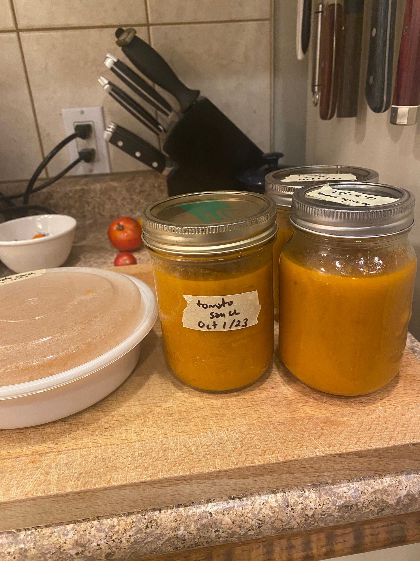 Three pint jars of orangeish tomato sauce, with masking tape labels showing the date. There is also a round white plastic container of the same. All of them rest on a wooden cutting board with a knife block in behind.