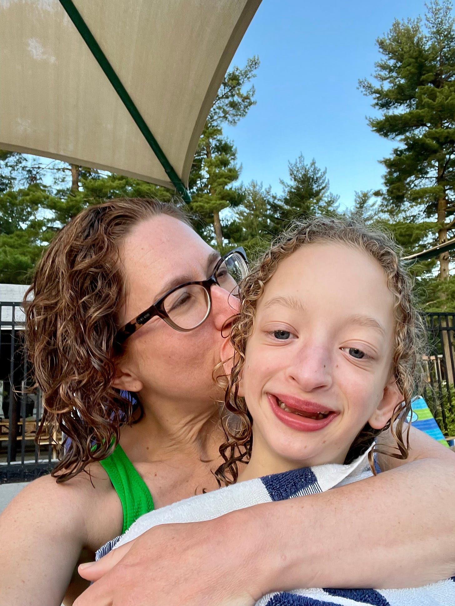 Selfie of my kid and me. My arm is wrapped around her. We both have wet hair. I'm kissing her head. She is in the foreground. She's wrapped in a towel. A beach umbrella and blue sky is above us.