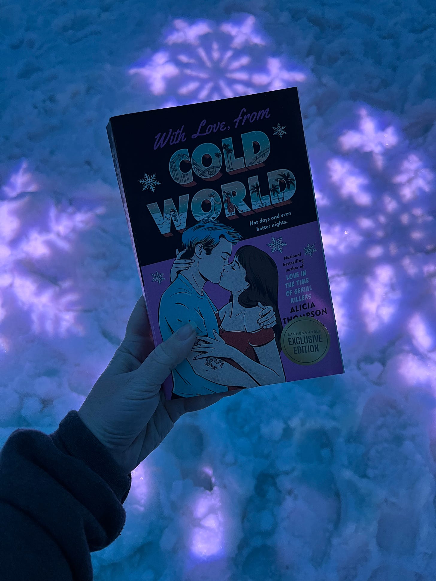 Picture of my hand holding up the pink cover copy of WITH LOVE, FROM COLD WORLD in front of the snow in the Igloo, which is lit by purple lights and has images of snowflakes projected on it