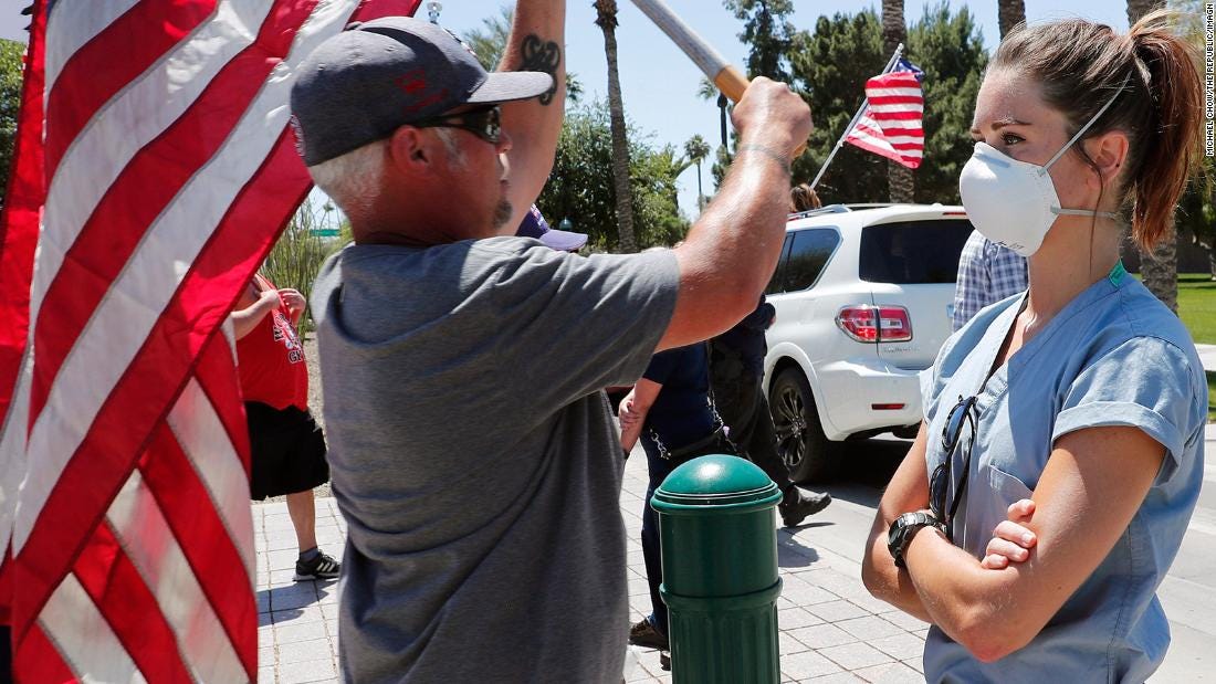 Man aggressively waving U.S. flag in the face of a healthcare worker who stands with mask on and arms crossed