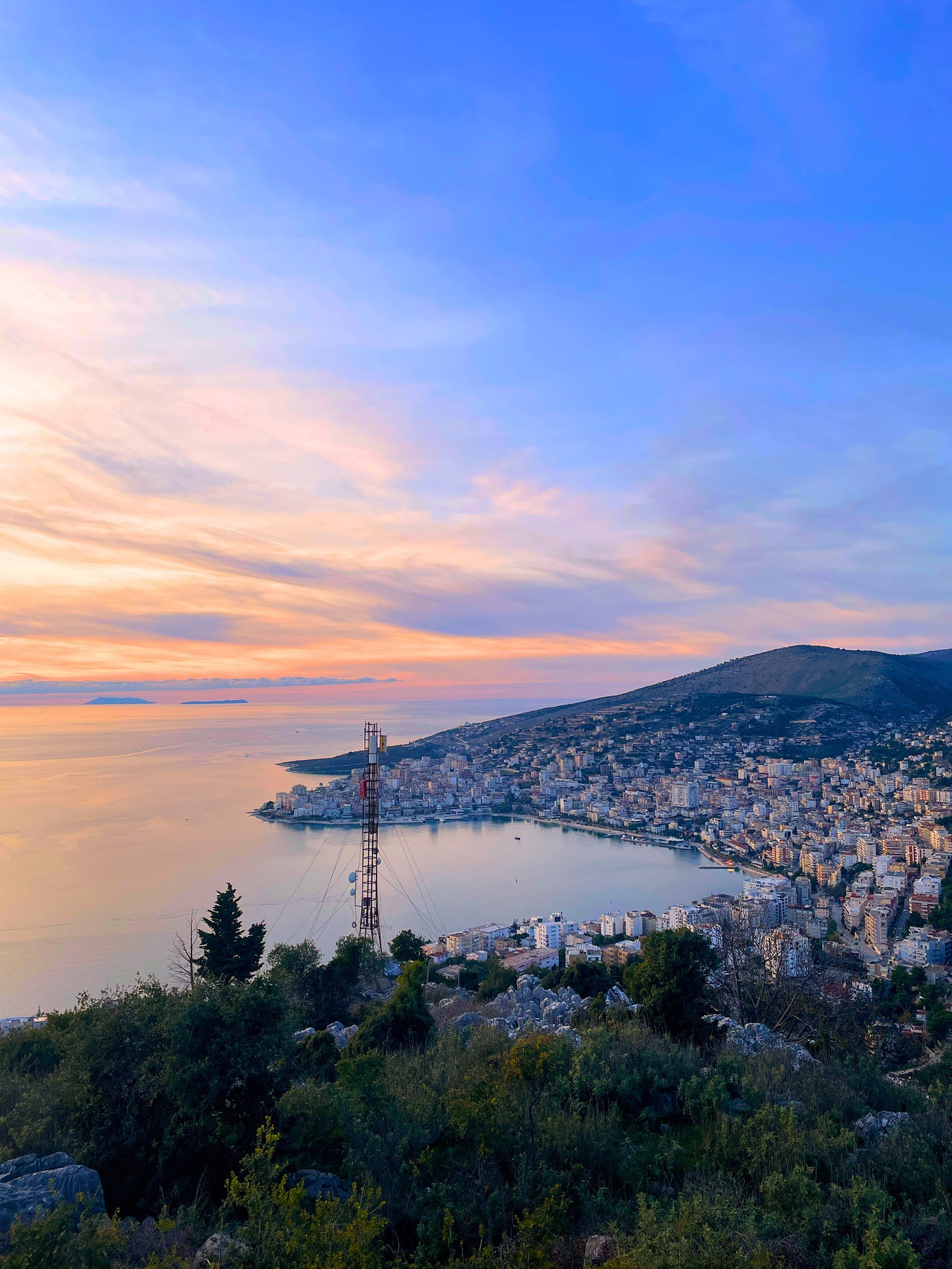 Foto do topo de uma montanha mostra a cidade, vários prédios, uma montanha, o horizonte com o céu azul, pôr-do-sol entre amarelo e rosa e montanhas em volta.