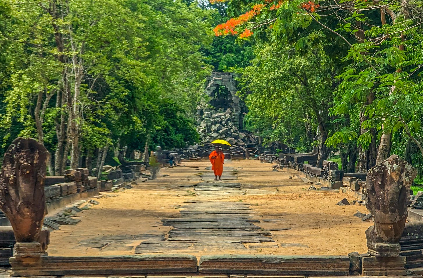 Photo of a ruined plaza in an Angkor Wat temple. A long entryway is surrounded by trees, a monk wearing an orange robe and holding a yellow umbrella is in the distance walking toward the camera. 