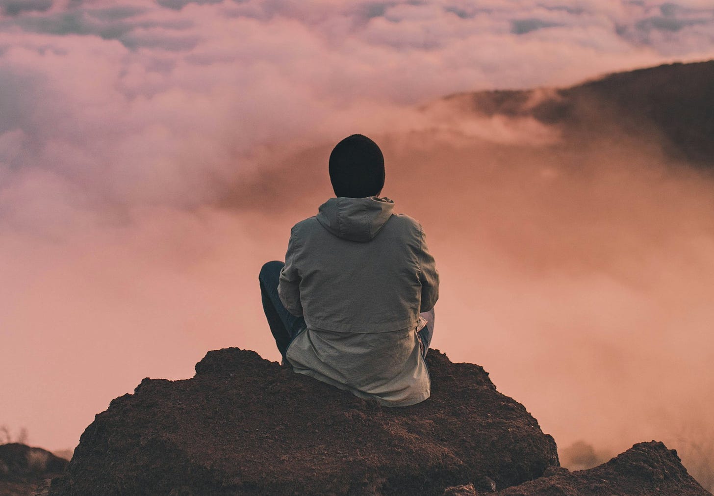 A man sits atop Mauna Kea, Hawaii, with pink clouds in front of him.