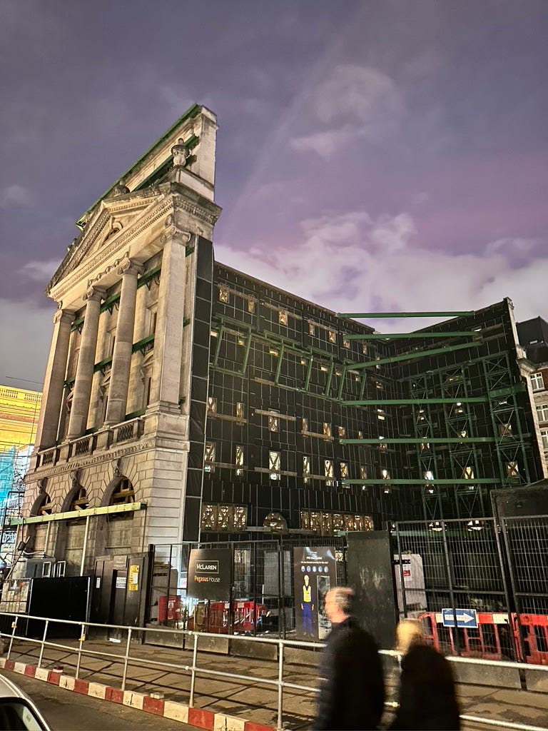 Partly demolished building with a preserved neo-classical front, taken on a dark evening with two passers by in the foreground.