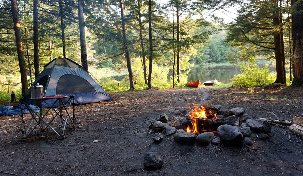 A campsite with a tent, table, coffee pot, canoe and a campfire