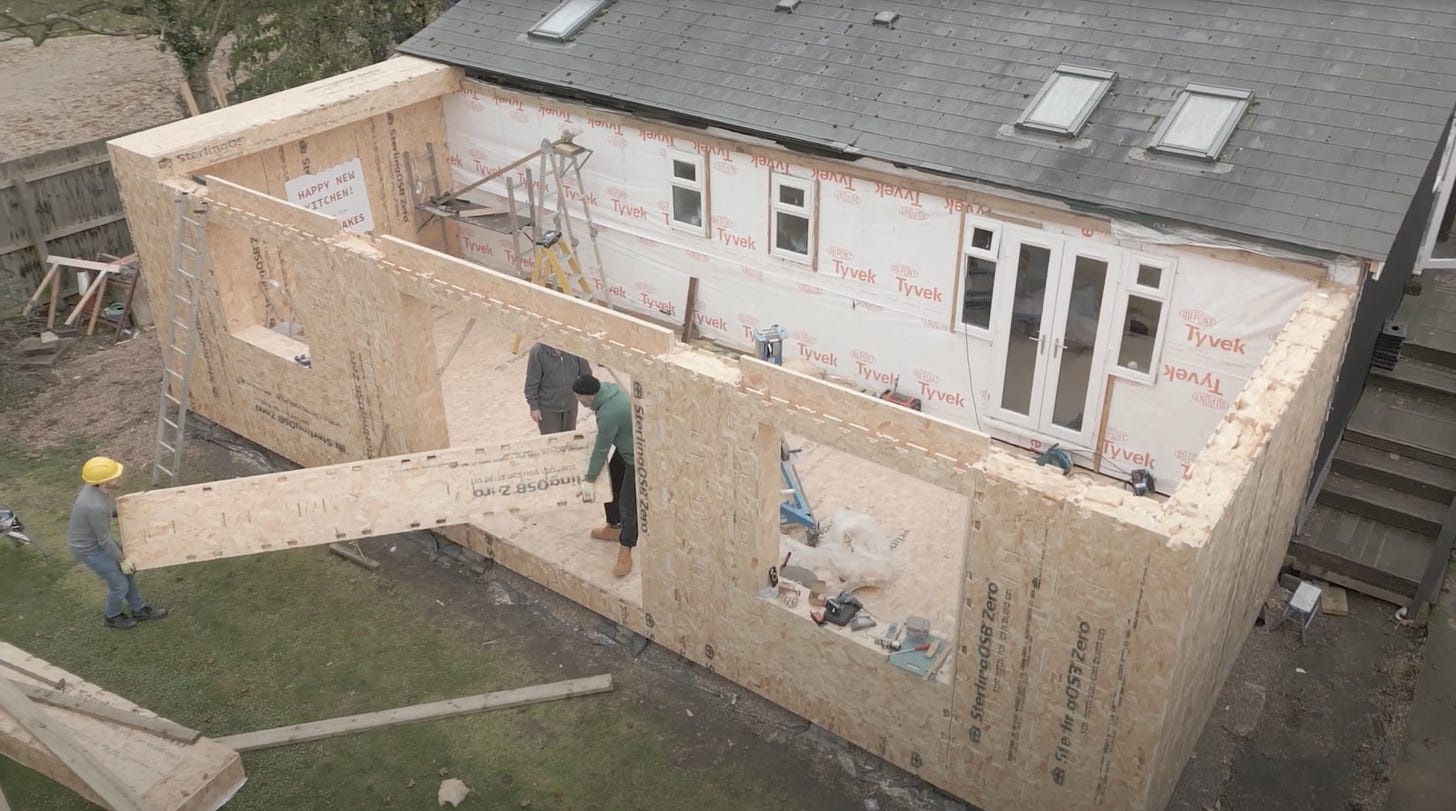 2 people carry a long WikiHouse block through a door opening into an extension. The extension is attached to another building and has a floor and walls but no roof.