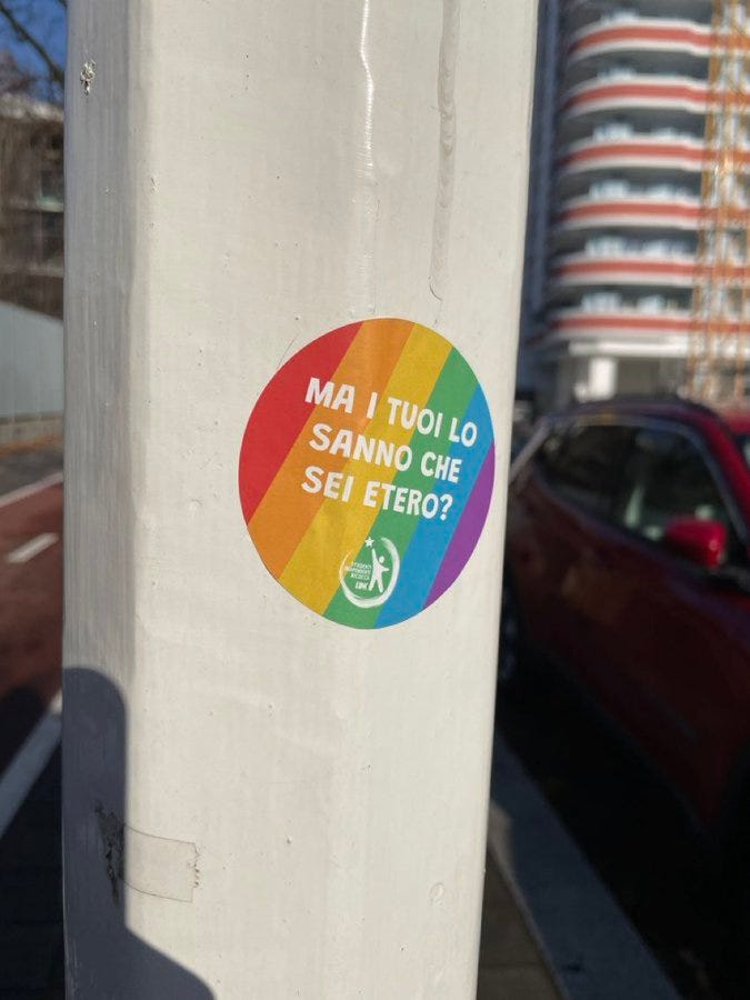 Sticker dai colori arcobaleno attaccato a un palo. La scritta al centro dice "Ma i tuoi lo sanno che sei etero?"