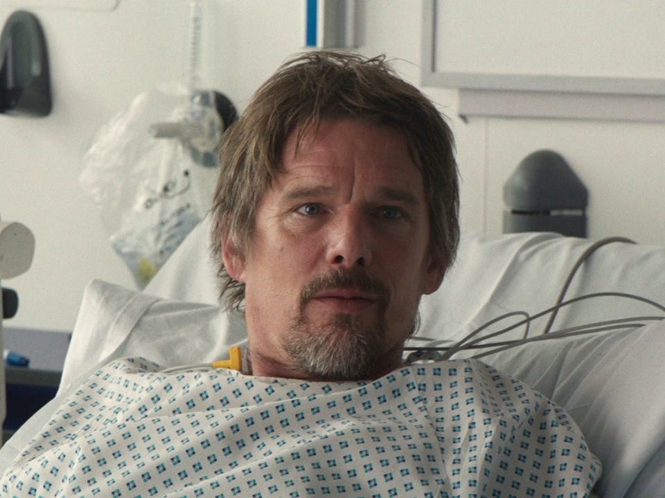 Still from Juliet Naked that shows Ethan Hawke in a hospital gown, lying in a hospital bed, and looking up at Annie (who we don't see in this picture). This is a man falling in love.