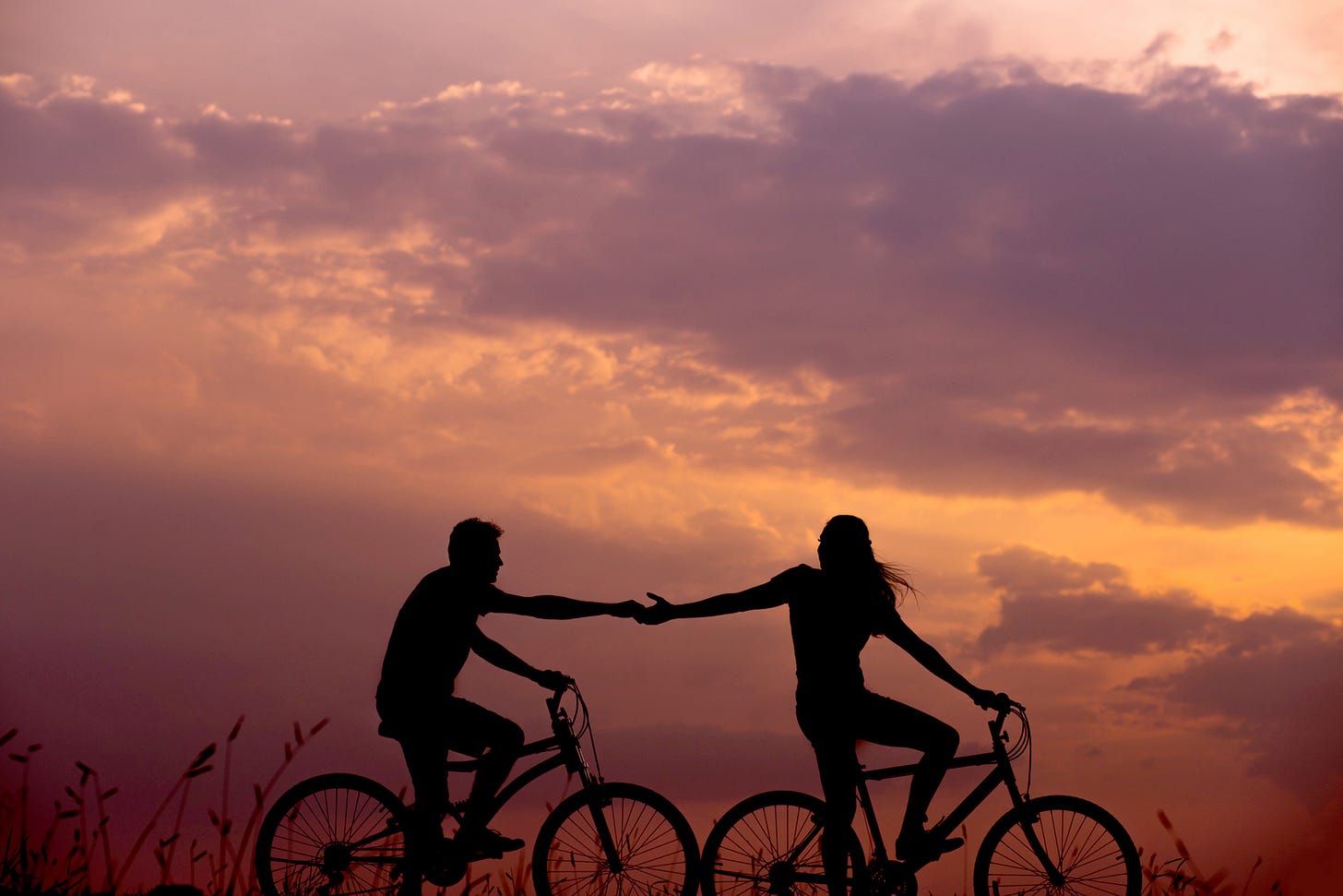 Silhouettes of a man and woman on bicycles with arms outstreched holding hands against a backdrop of a pink and purple cloudy sky.