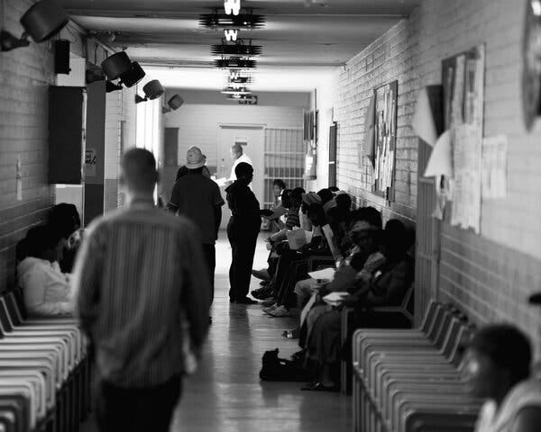 A view down a brick-walled corridor lined with people sitting in chairs. 