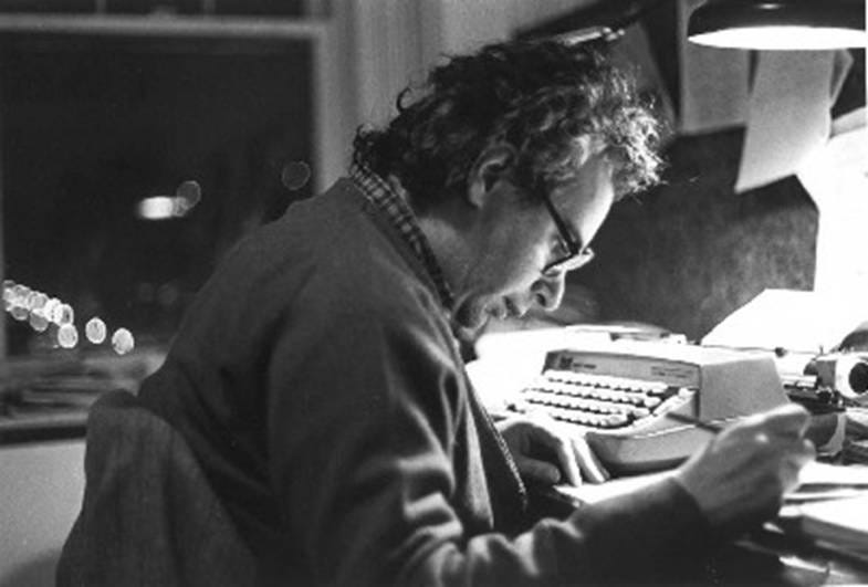 In a black-and-white photo, a bespectacled man with unruly hair sits at a desk, holding a pen or pencil over a manuscript. On the desk is a manual typewriter with a sheet of paper in the spool. Outside a window, we see what looks like a sequence of blurry headlights in the night.