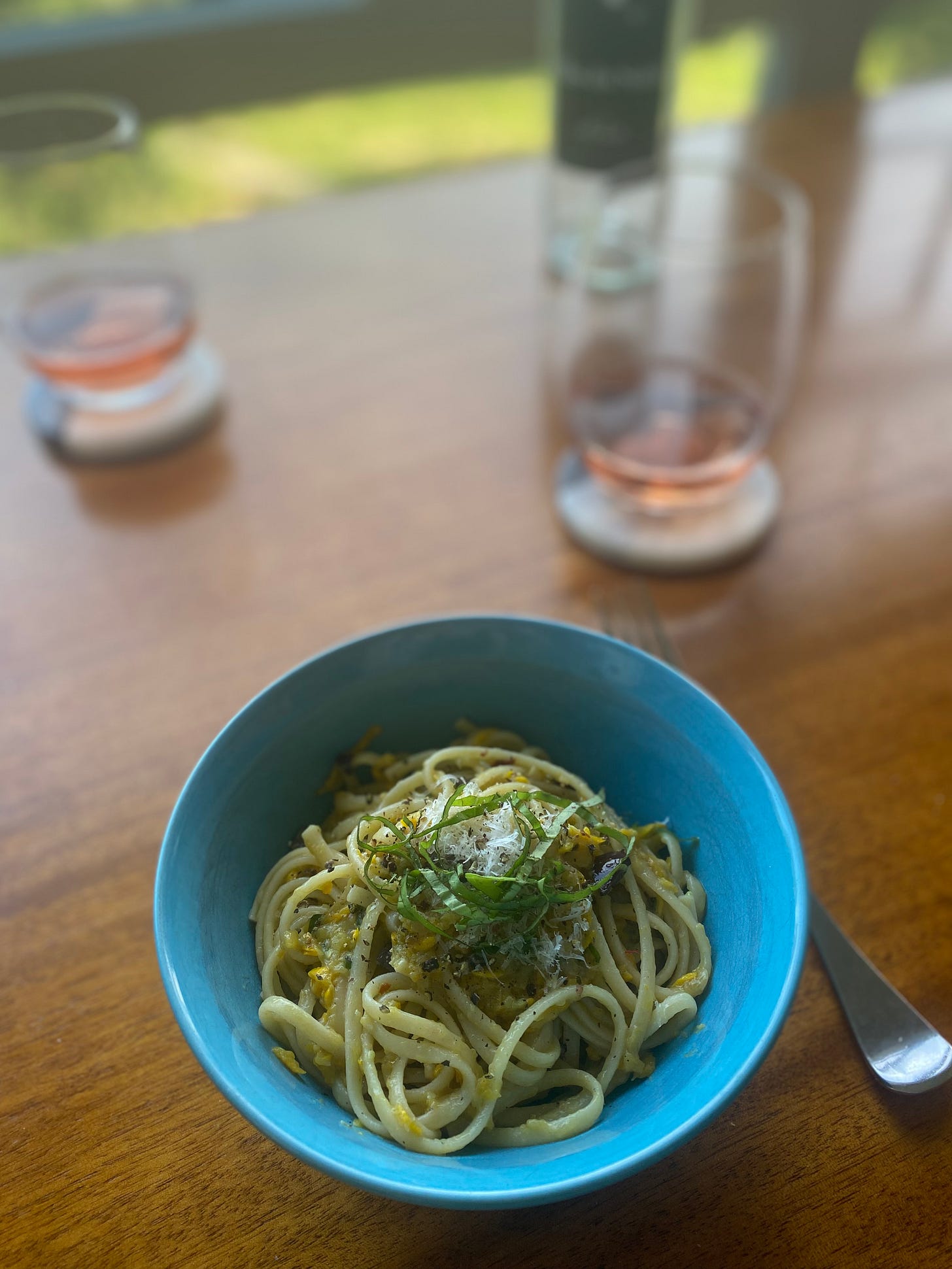 In a blue bowl, the zucchini butter pasta described above, made with yellow zucchini and kalamata olives. On top is are basil, parmesan, and ground pepper. Two coasters in the background have glasses of rosé wine on them.