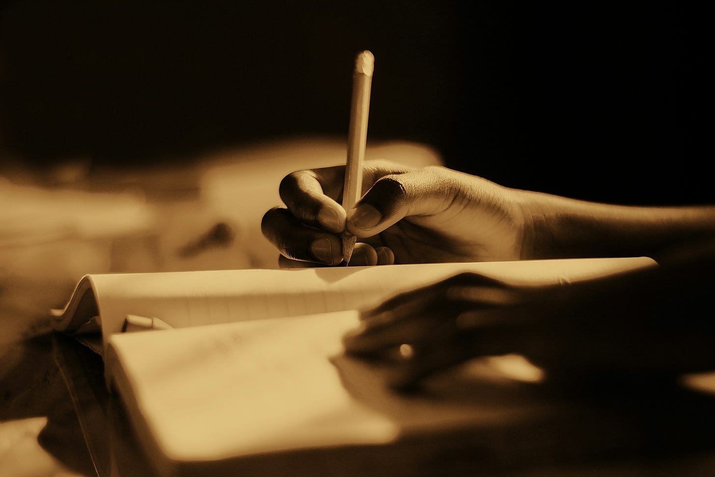 Toni seppia con una mano destra che spunta da destra e con una matita scrive qualcosa su un libro aperto, mentre l'altra in basso riposa distesa sulle pagine. Sfondo nero e sfuocato.