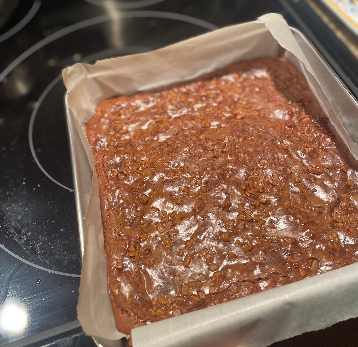 A square baking pan lined with parchment and filled with unsliced brownies, topped with small toffee pieces which nearly match the brownies in colour. 