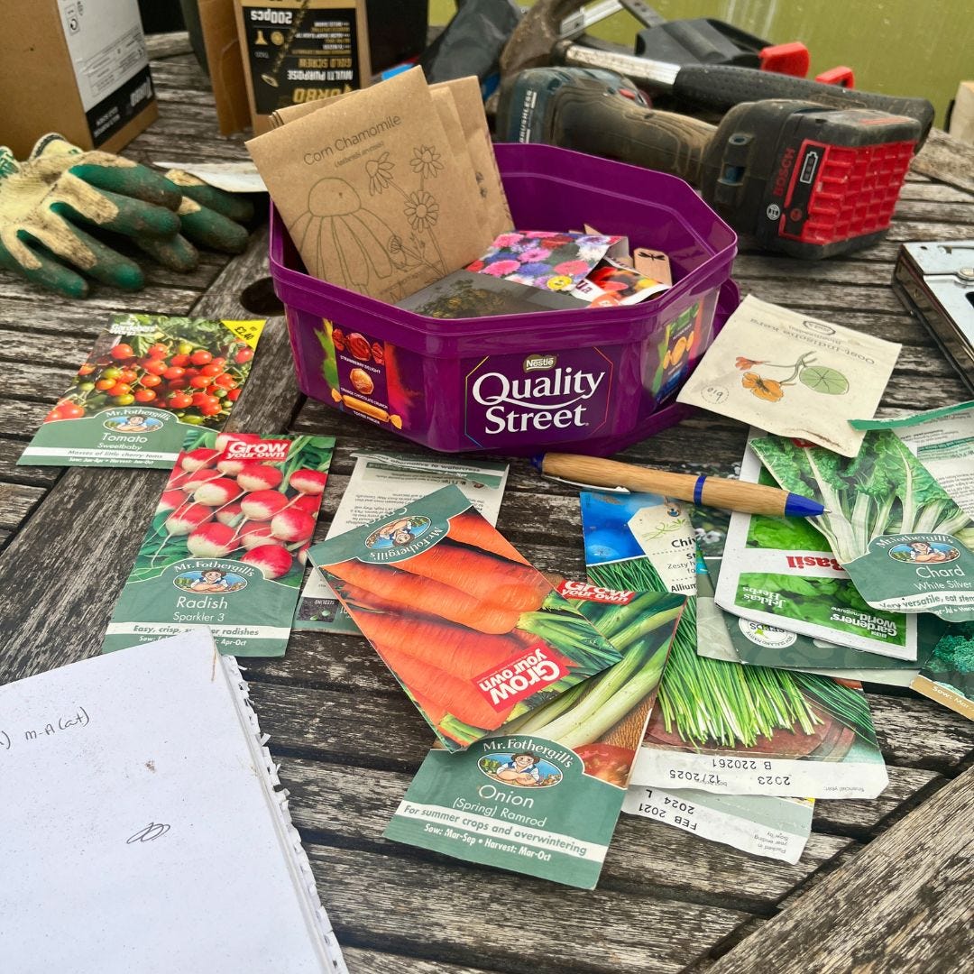 A table with gloves & tools on, along with a quality street tub with packets of seeds & other packets of seeds strewn across the table.