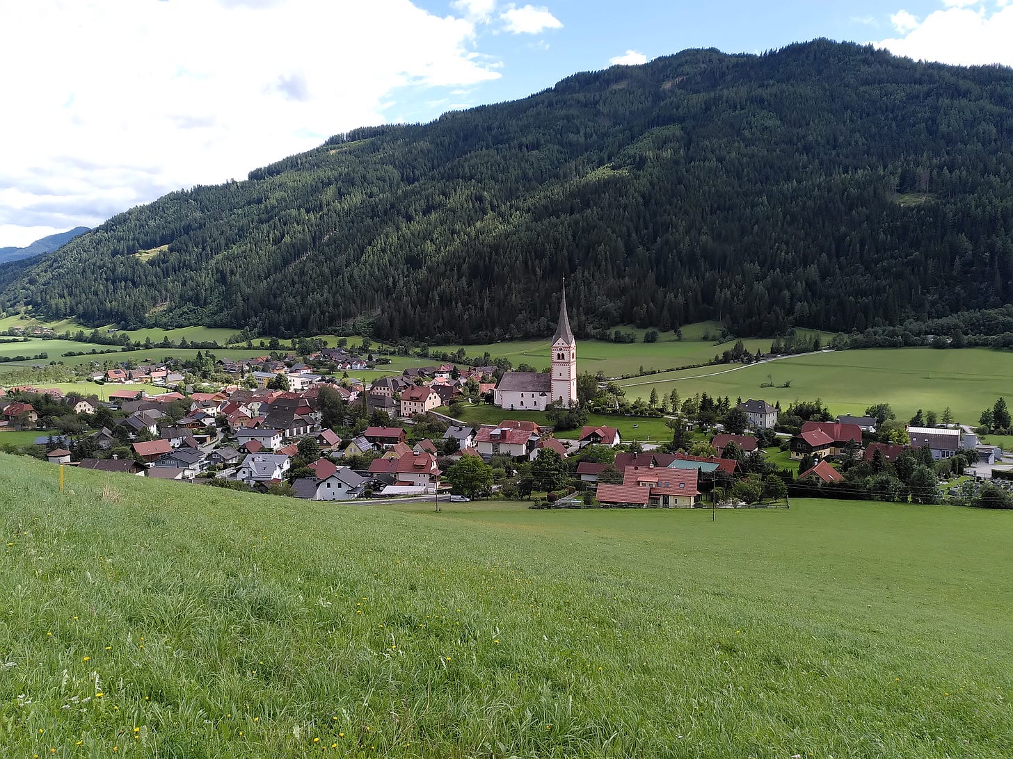 Central Austrian town nestled in Alpine hills, wooded hills in background; green grassy hills in foreground. Church spire and surrounding buildings visible from afar.