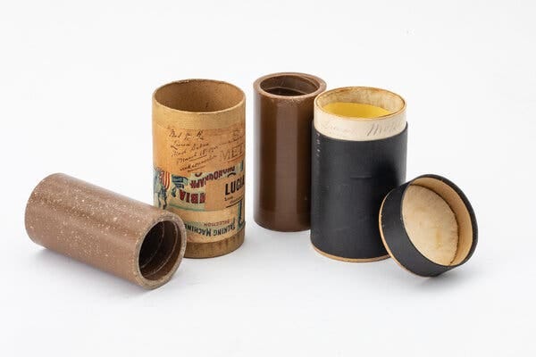 Wax cylinders and open cardboard canisters sit on a white background. One canister is black and the other is covered with labels and handwriting with words that include “talking machine” and “Met.”