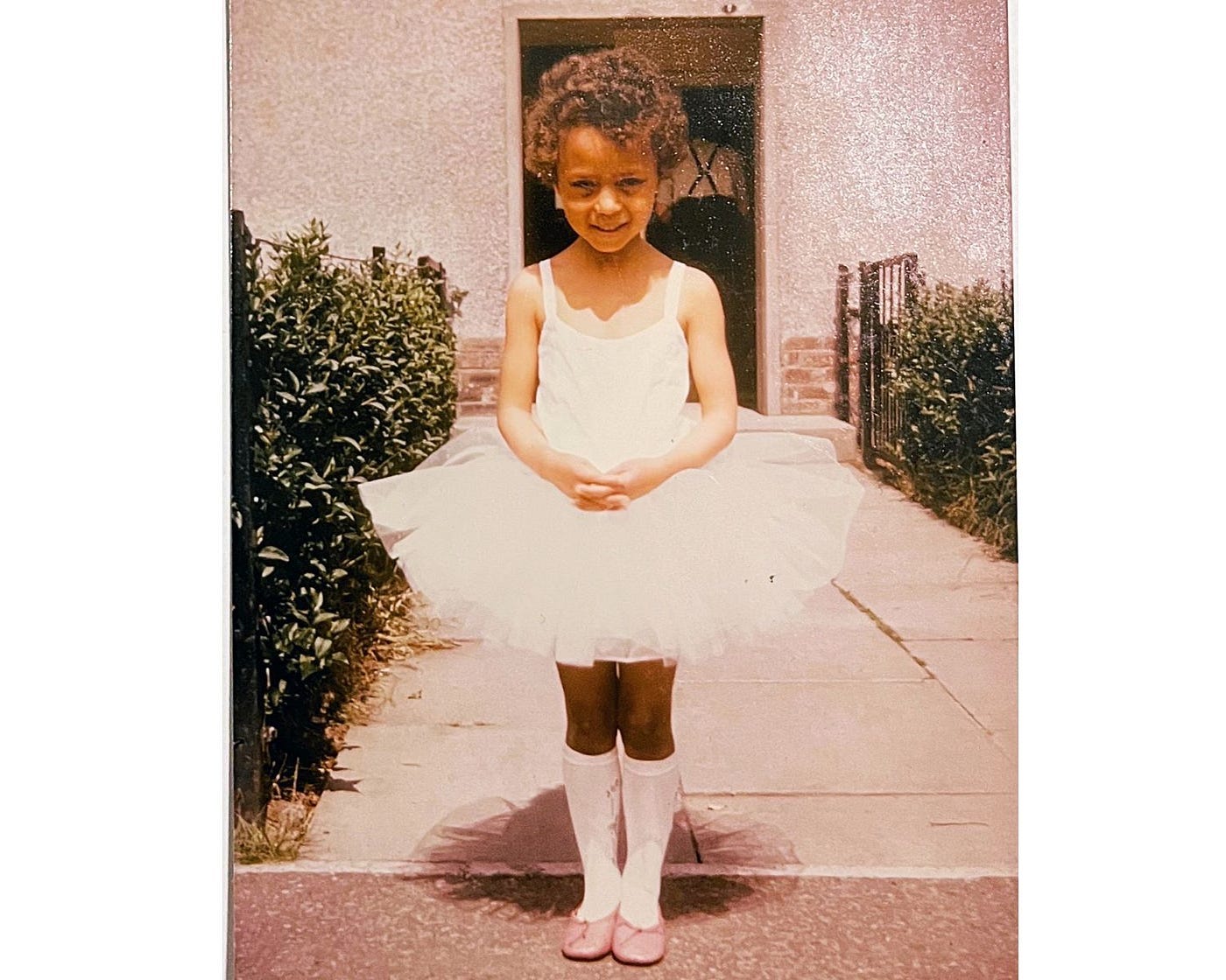 Me, about age 8, standing on a paved path leading to dark entry, waring a white tulle skirt, white knee socks, and pink ballet slippers