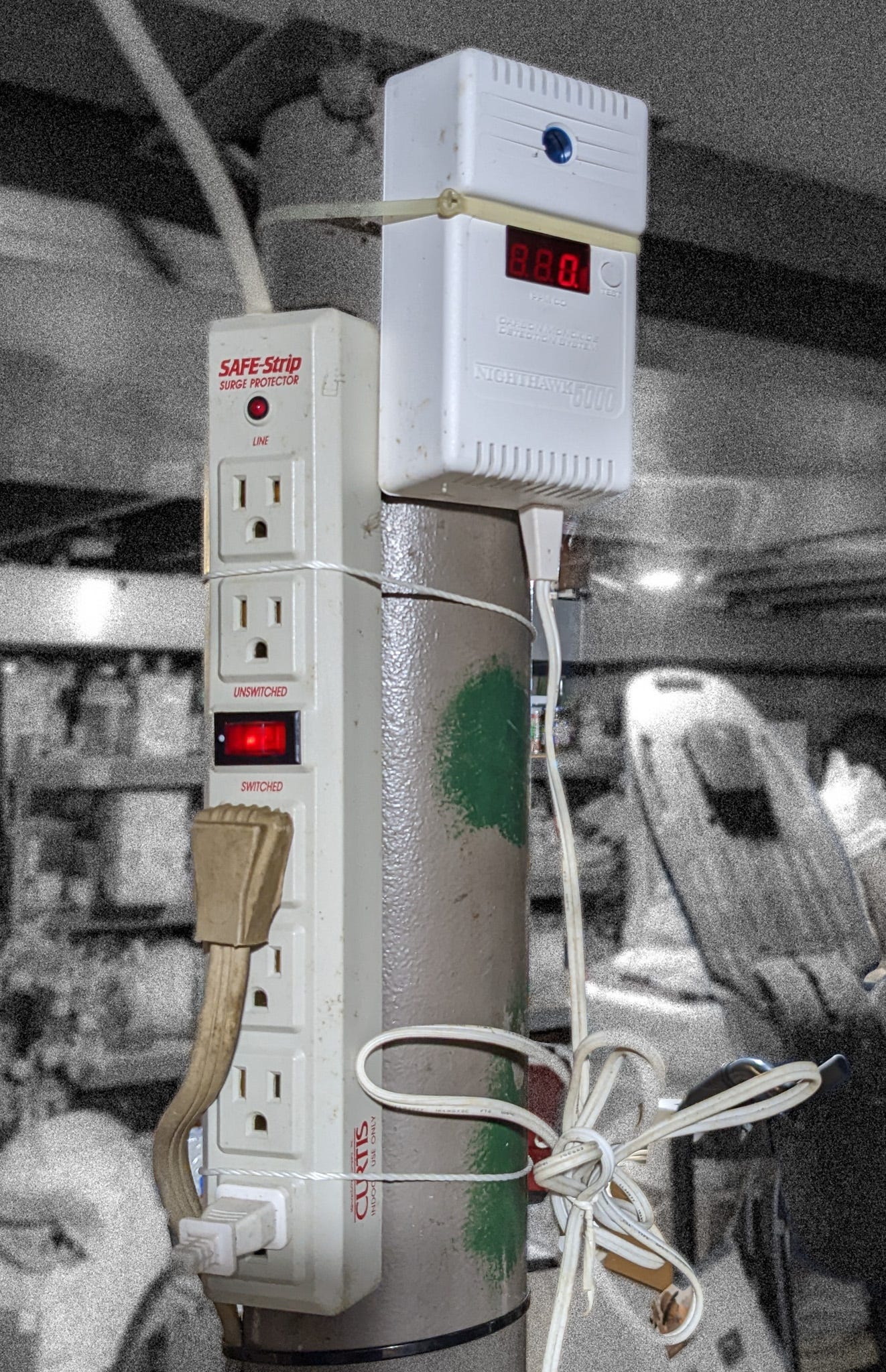 A photograph taken in a very cluttered basement. The focus is on a post -- a sturdy pipe -- between the floor and ceiling; to the post are affixed a surge protector and a programmable household security device. The surge protector has a couple of things plugged into it, and a glowing red rocker switch labeled (in red) "UNSWITCHED" and "SWITCHED."