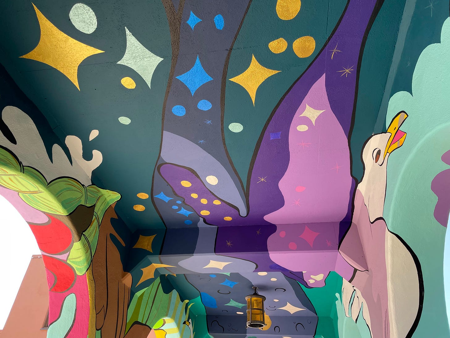 A mural, shown from below, covers a ceiling and pillars. It depicts a seagull, as well as various shapes and formations, using a mix of purple, yellow, white, red, green, blue, and pink. 