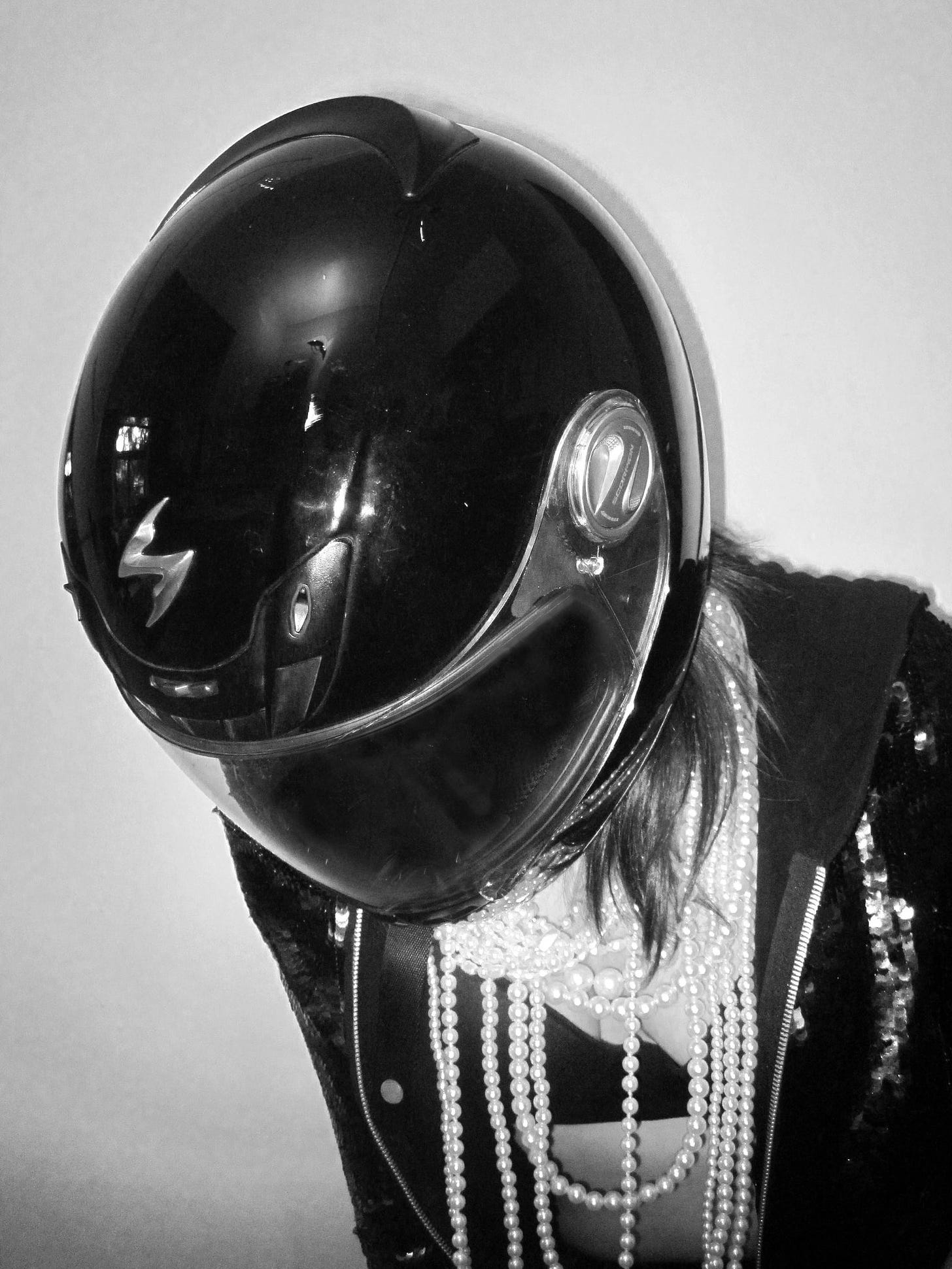 Image is a black and white photo of a person wearing a motorcycle helmet with piles of pearls dripping from their neck down across their chest down to their navel. They are wearing a sequined hoodie that is open, revealing a black bra beneath the pearls. They're standing in front of a stark, bare wall.