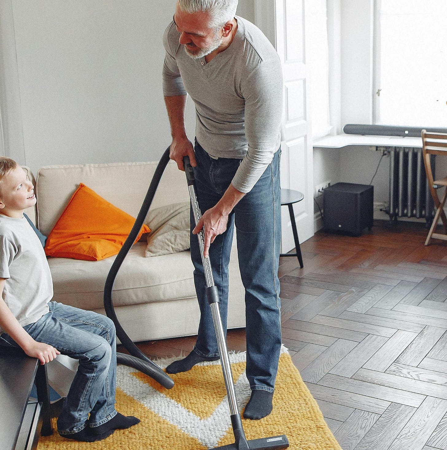 Man vacuuming rug while talking to seated boy