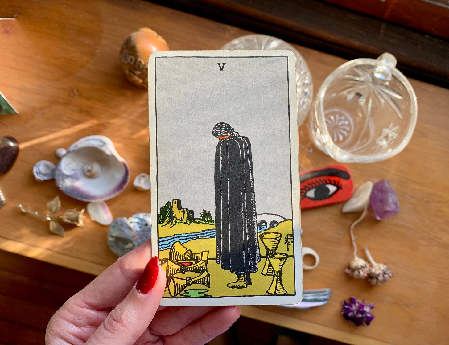 A hand is holding a Tarot card in front of a shelf with a bunch of random objects on it, shells, talismans, glass objects and dust. The card is Five of cups by Pamela Colman Smith for the Rider-Waite-Smith Tarot. In the image a person is dressed all in black, looking down at three spilled cups with two cups standing upright behind them. There is a castle in the distance across a river with a white bridge going over it.