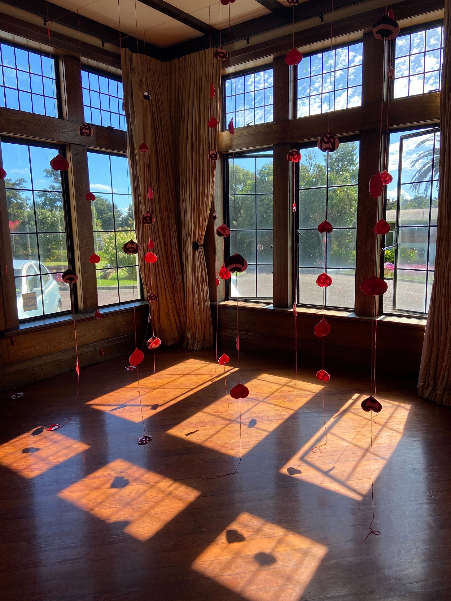 Strings of red hearts are draped from the ceiling in a wooden room and windows with the shadows reflecting on the floor.