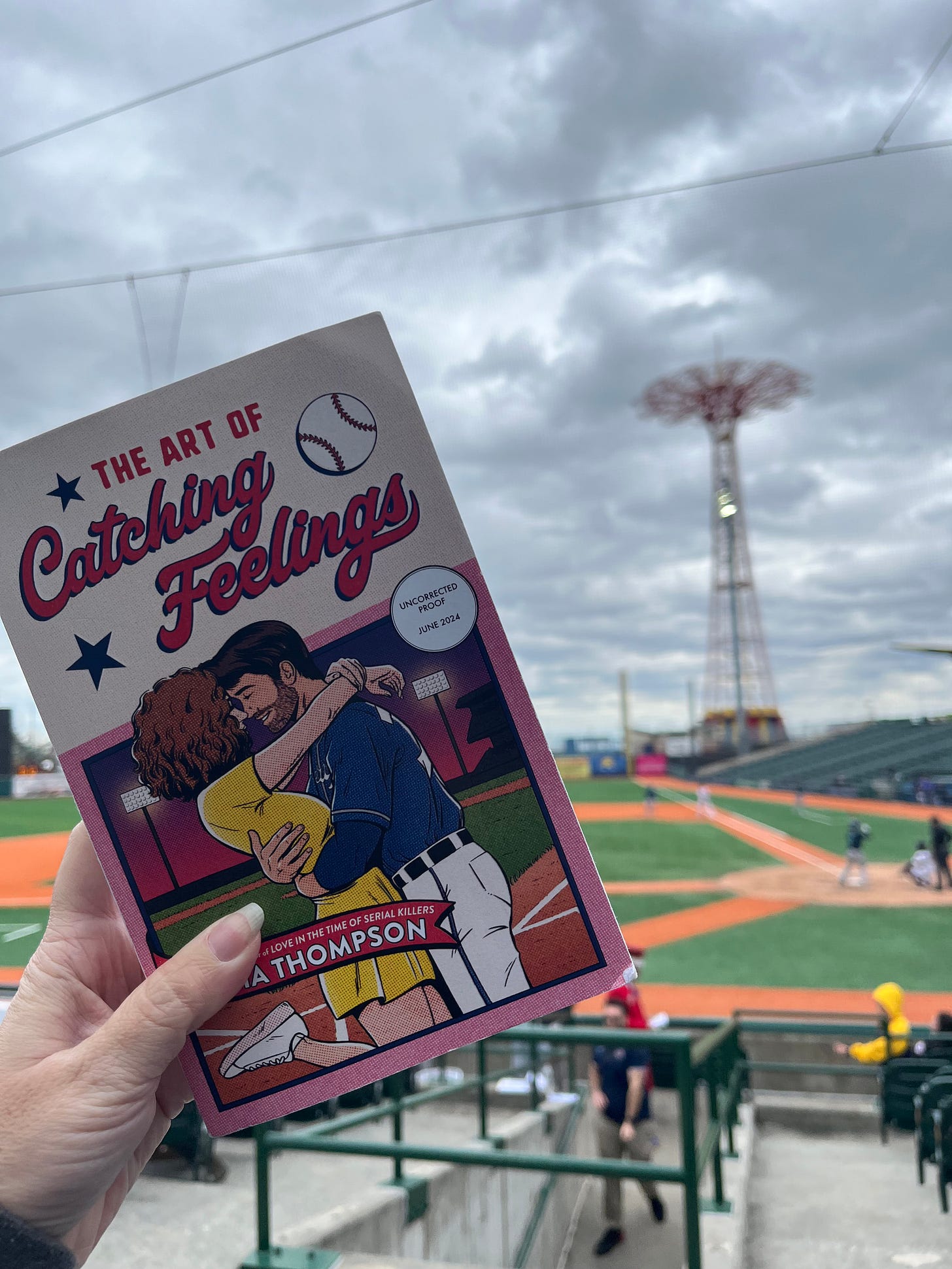 Picture of my hand holding up a copy of The Art of Catching Feelings in front of the Brooklyn Cyclones Game