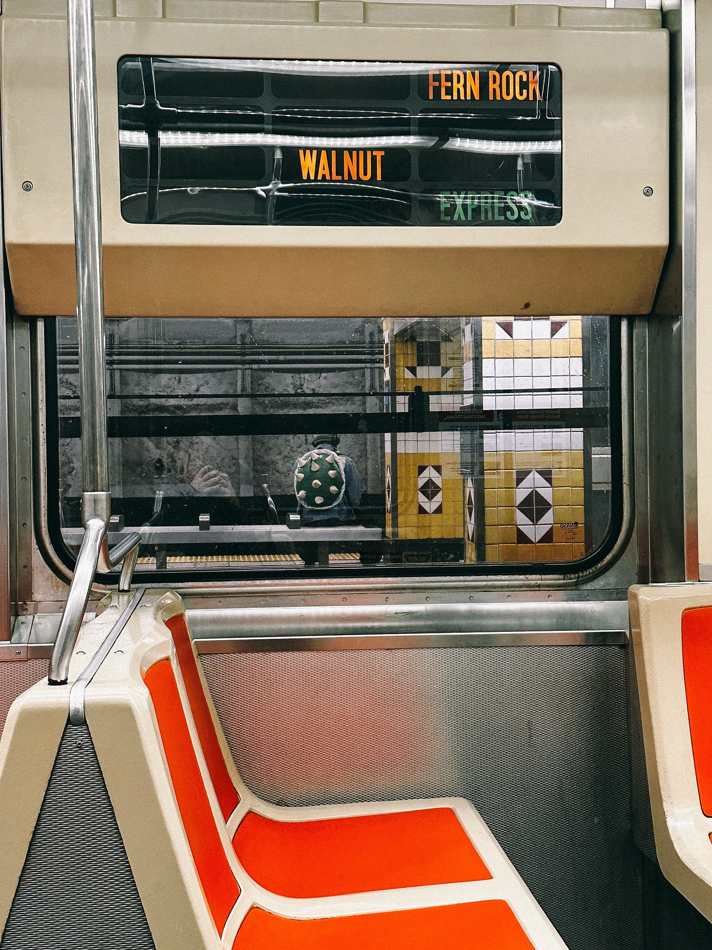 view from inside a train car, orange seats in the foreground and a sign at the top that says FERN ROCK WALNUT EXPRESS. out the window someone wearing a Bowser backpack is visible sitting on a bench at a station.