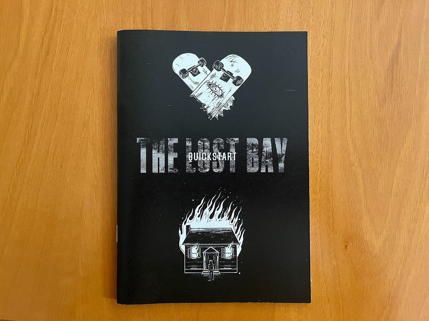 A black zine reading The Lost Bay Quickstart. Above the text is a broken skateboard forming a heart, and below is a burning house.