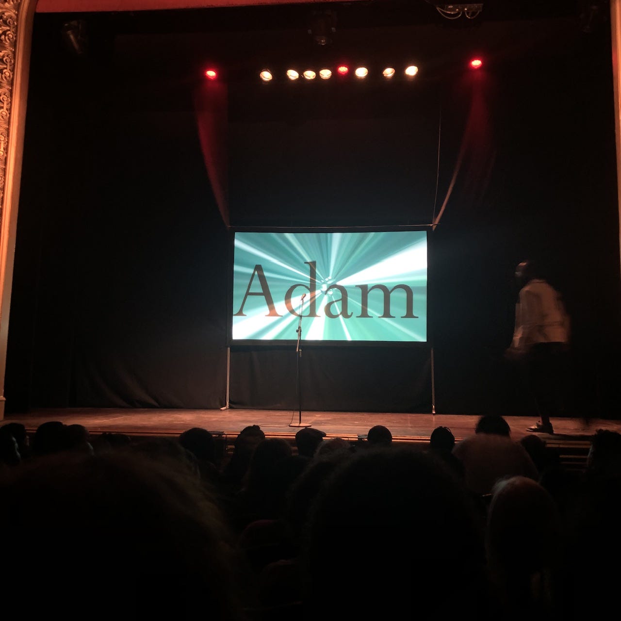 Stage of Stratford East theatre with "Adam" projected on a screen and a reader walking up to a microphone