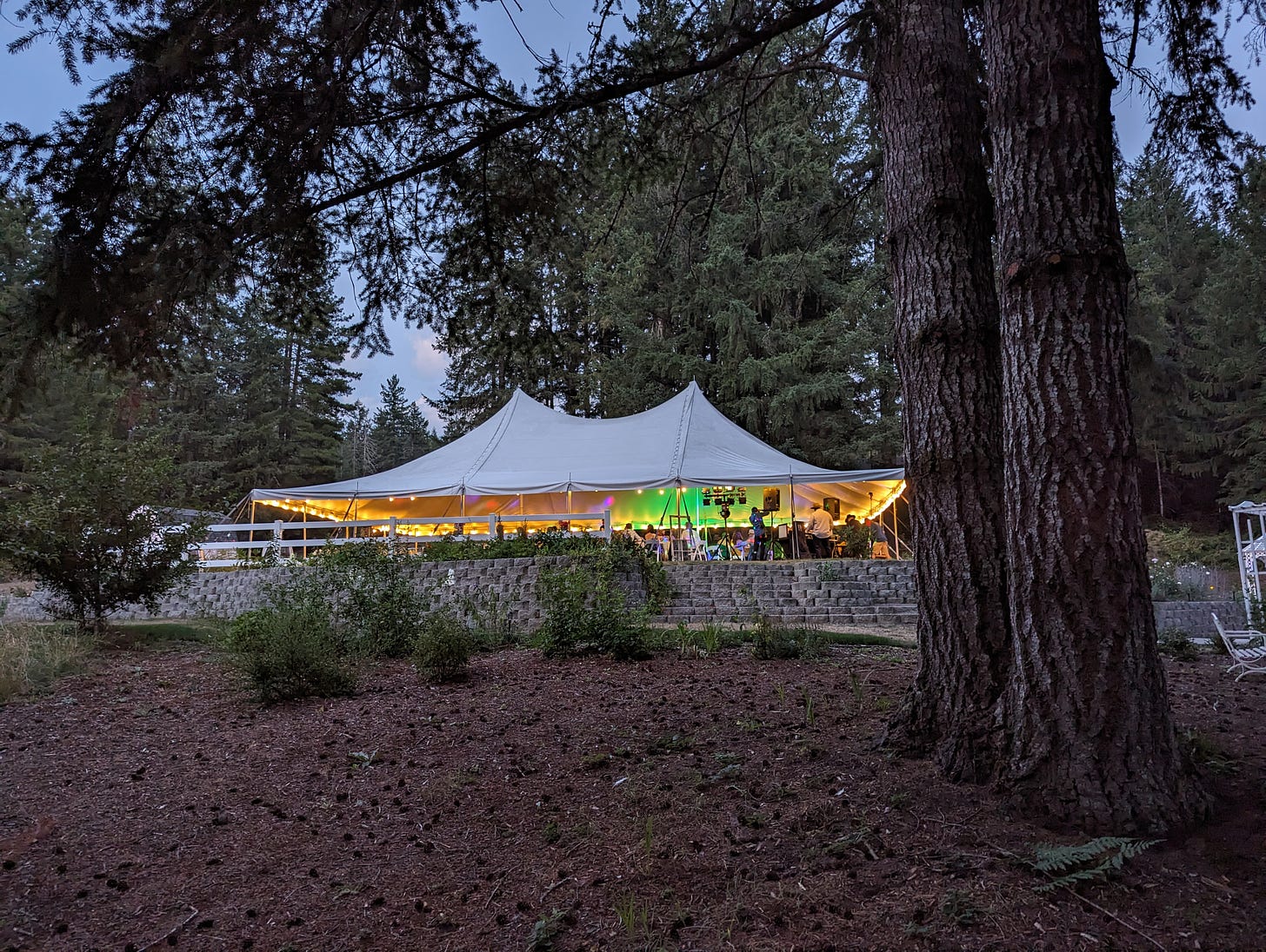 A brightly lit massive white pavilion tent in the evening, filled with dancing people and bright colorful lights.