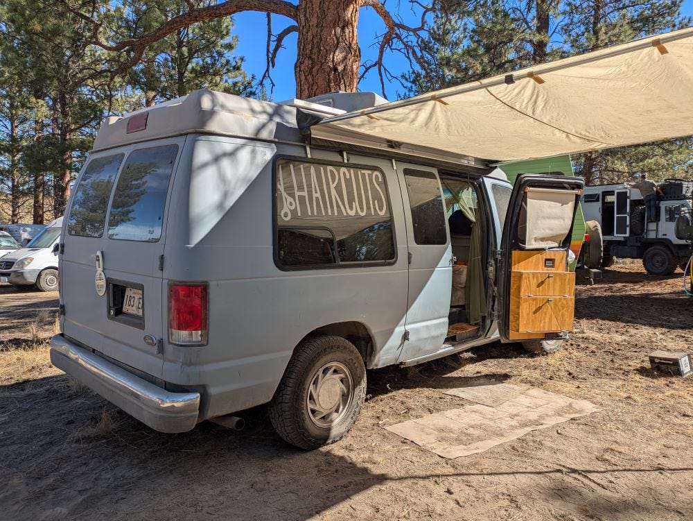 a blue van with a sign for haircuts in the window, set in a dirt campground surrounded by other vehicles under blue sky and pine trees