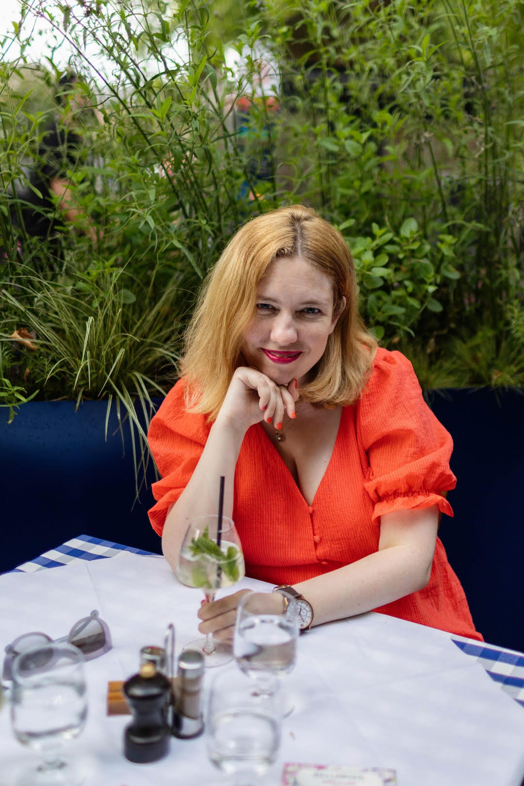 Sian is a white woman with shoulder-length, auburn hair, sitting at a table in a garden and wearing a coral coloured dress.