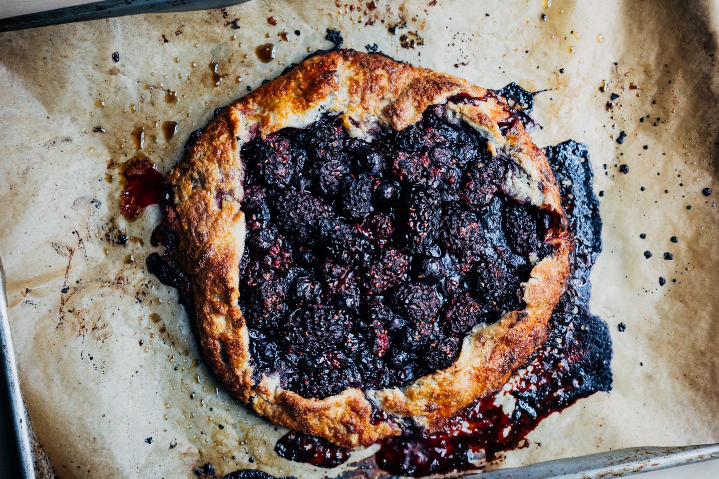 A baked berry galette on a baking sheet with bit of caramelized and burnt filling around it. 