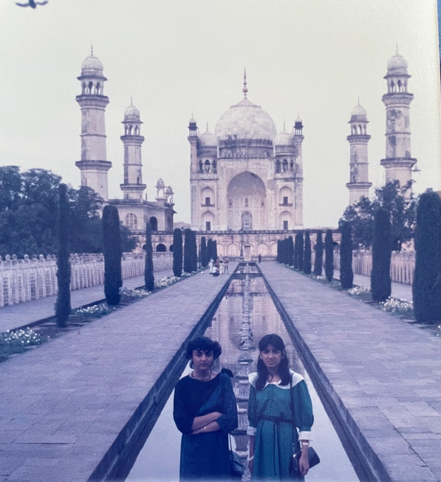 Two teenage Indian women wear dark dresses and stand in front of the Taj Mahal