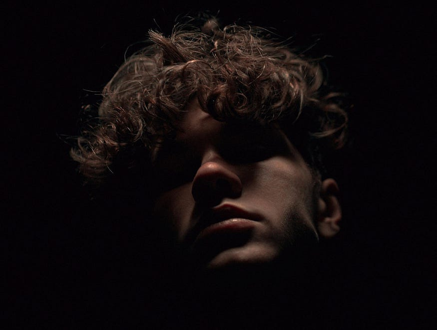 A man’s face is visible amid a black backdrop. He has curly brown hair. A man’s face is visible amid a black backdrop. He has curly brown hair.