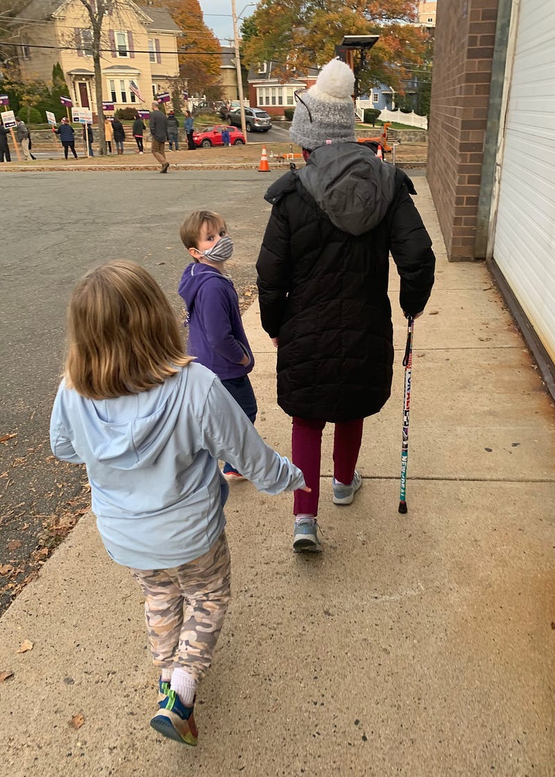 The author (white woman in black coat) walking with a cane — pictured from behind, with two children next to her.