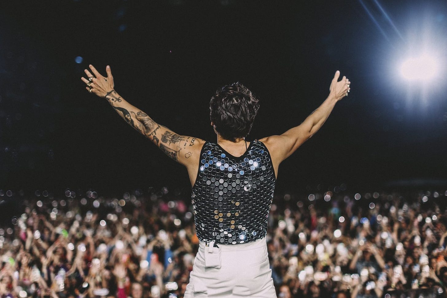 Harry Styles on stage, seen from the back, wearing white pantss and a silver sequinned top, his arms outstretched. The crowd is in the background, out of focus,