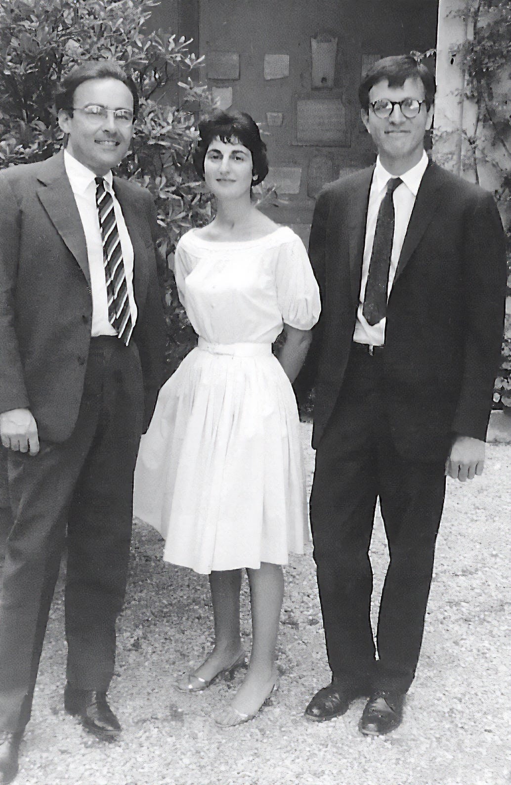 A young woman in a cocktail dress stands smiling flanked by two men in suits and ties wearing glasses. They stand in a courtyard with vines and trees behind.