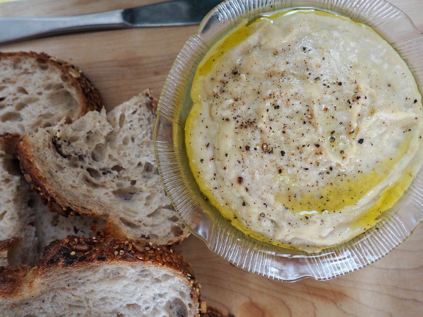 A bowl of white bean dip drizzled with extra olive oil and sprinkled with salt and pepper. To the left of the bowl there are slices of seedy bread.