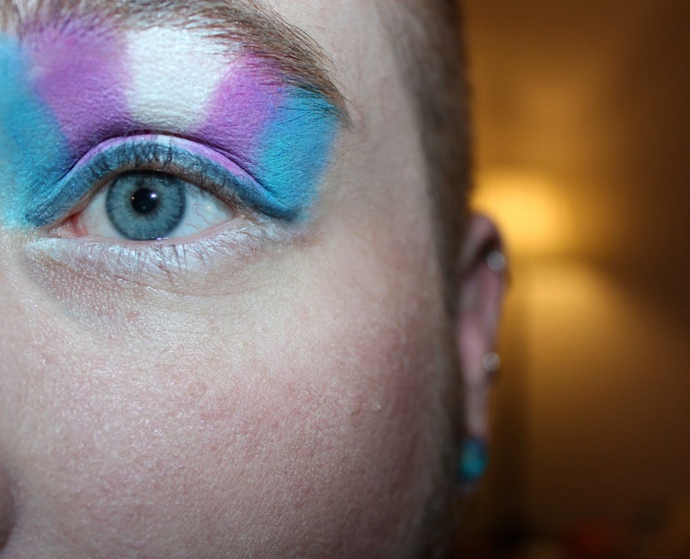 Photo of a white person with blue eyes and bright blue, pink, and white eyeshadow like the colors of the trans flag